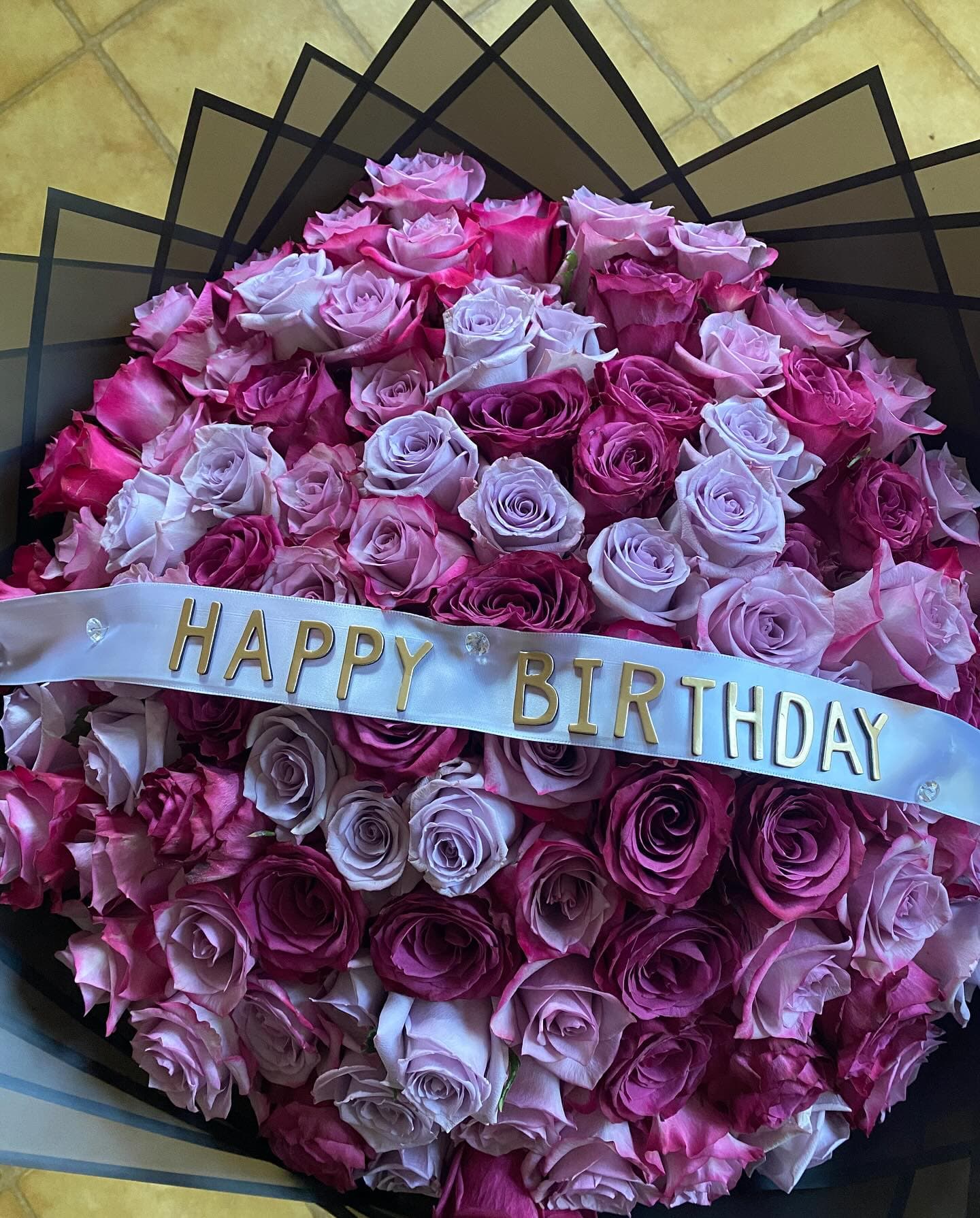 Large bouquet of purple and pink roses with a white 'HAPPY BIRTHDAY' ribbon in gold.