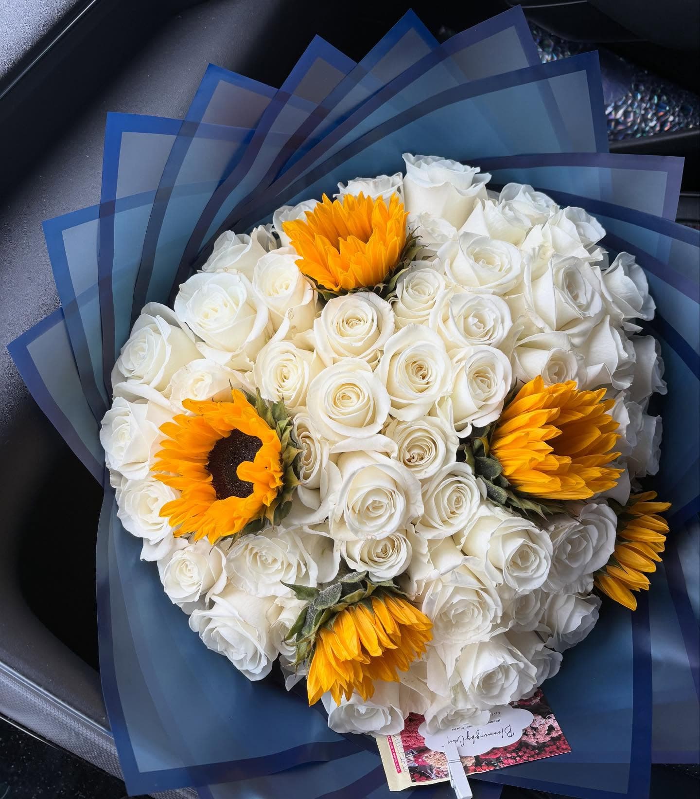Large bouquet of white roses and yellow sunflowers wrapped in elegant blue translucent paper.
