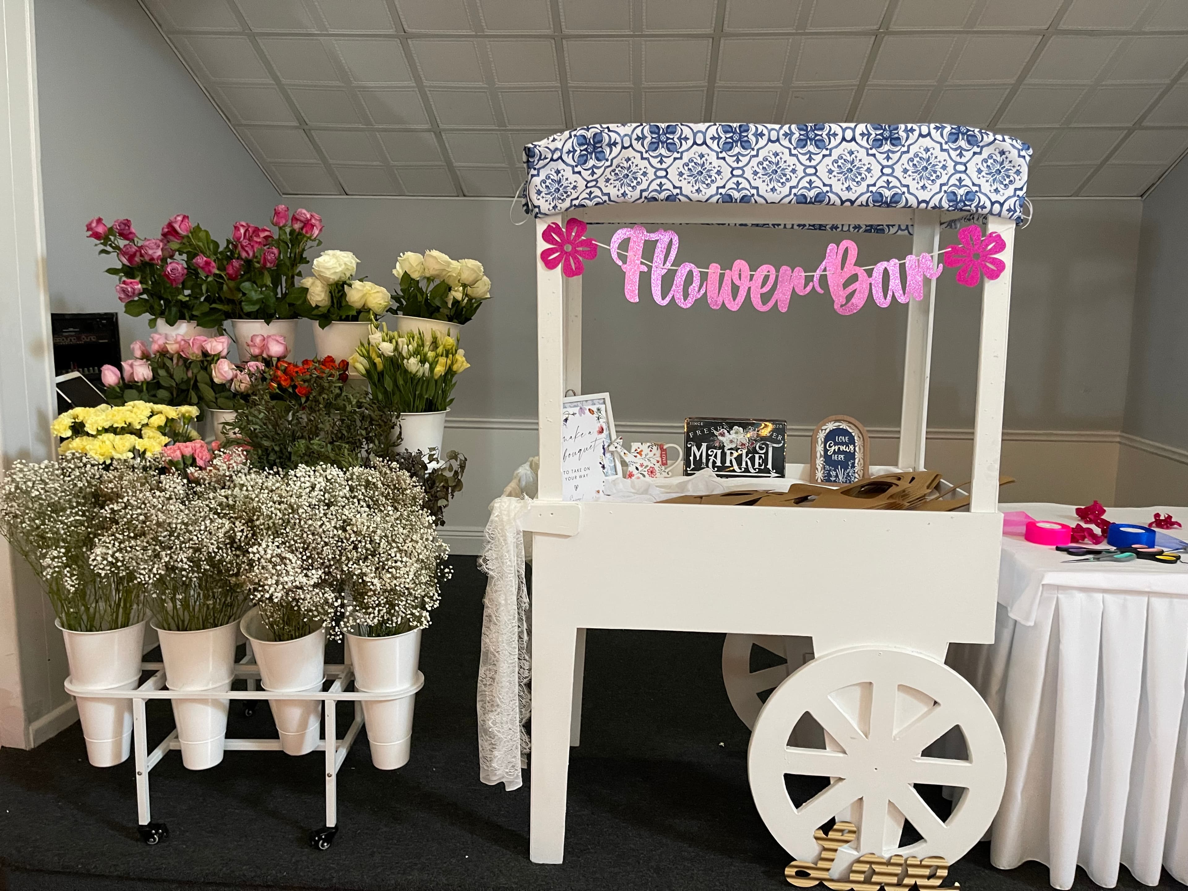 White wooden flower bar cart with pink banner and buckets of colorful assorted flowers.
